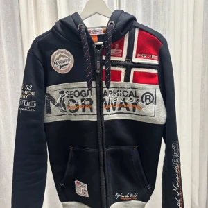 Geographical Norway hoodie 🤩 - Hoodie från Geographical Norway med broderade och tryckta detaljer, norsk flagga på bröstet och patchar på ärm och bröst. Grå och röda inslag, dragkedja framtill, huva med snörning och två fickor. 