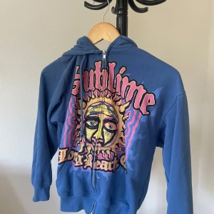 Blå Sublime hoodie med tryck - Högst längd: 170cm. Om du är längre än så kommer den vara för liten. Säljer en blå hoodie från Divided med stort Sublime-tryck i färgglada toner på framsidan. Tröjan har huva med dragsko, lång ärm och hel dragkedja framtill. Perfekt för dig som gillar bandmerch och streetstyle.