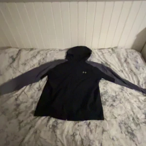 Svart vindjacka från Under Armour - Svart vindjacka från Under Armour med gråa ärmar och klassisk logga på bröstet. Tröjan har huva och långa ärmar, perfekt för träning eller chill. Materialet känns sportigt och lätt, och passformen är normal. Snygg och enkel stil för dig som gillar sportiga vibes.