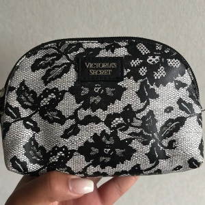 Svartvit blommig necessär Victoria's Secret - Snygg liten necessär från Victoria's Secret i svart och vitt med blommigt spetsmönster. Väskan har dragkedja upptill och är perfekt för smink eller småprylar. Stilren och trendig design som sticker ut i väskan eller på resan.
