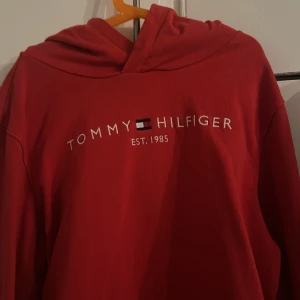 Röd hoodie från Tommy Hilfiger - Röd hoodie från Tommy Hilfiger med vit broderad logga och text på bröstet. Klassisk huva och långa ärmar, perfekt för en chill och stilren look. Mjuk bomullskänsla och enkel att matcha med jeans eller joggers.