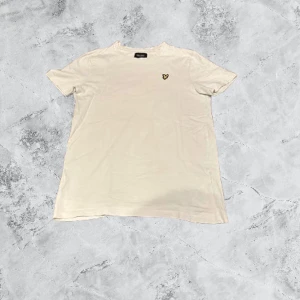 Vit t-shirt från Lyle & Scott - Vit t-shirt från Lyle & Scott med klassisk rund hals och liten gul logga på bröstet. Enkel och stilren design som passar till det mesta. Tillverkad i mjuk bomull för en skön känsla hela dagen.
