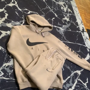 Beige Nike hoodie med svart logga - Säljer en beige hoodie från Nike med stor svart Swoosh och AIR-tryck på bröstet. Tröjan har huva med snörning och tryck längs ena ärmen. Perfekt för dig som gillar streetwear och sportig stil.