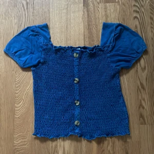 Blå blus med knappar från Zara Kids - Supersöt blå blus från Zara Kids med puffiga korta ärmar och smockad kropp. Framsidan har fyra stora knappar för design och blusen har en lätt croppad passform. Perfekt för dig som vill ha en söt bekväm tröja!