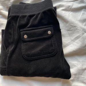 Svarta mjukisbyxor Juicy Couture, storlek XS - Svarta mjukisbyxor från Juicy Couture i mjuk velour med bred resår i midjan och snörning. Byxorna har fickor fram och bak med lock och silverfärgade knappar samt broderad JC-logga framtill. Perfekta för en chill och avslappnad stil.