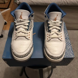 Nike Air Jordan 3 Retro White/Blue - Nike Air Jordan 3 Retro i vitt skinn med blå och kopparfärgade detaljer. Klassisk elephant print runt tå och häl, blå innersula och snörning. Snygg siluett med hög komfort och ikonisk Jumpman-logga på plösen. Perfekta för dig som vill sticka ut med en tidlös sneaker.