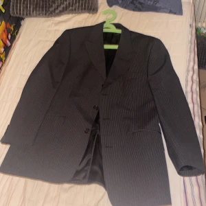 Svart randig kostym från Carlo Monti - Snygg svart kostym med diskreta vita ränder från Carlo Monti. Setet består av en klassisk kavaj med två knappar och en matchande väst med fem knappar. Båda plaggen har en stilren passform och är fodrade med svart tyg. Perfekt för dig som vill ha en clean och sofistikerad look.