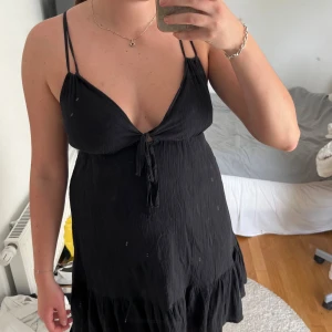 Svart kort klänning med knyt - Säljer en svart kort klänning med tunna axelband som korsas i ryggen och v-ringning med knyt framtill. Klänningen har volang nertill och är tillverkad i ett lätt, skrynkligt tyg som känns luftigt och skönt.