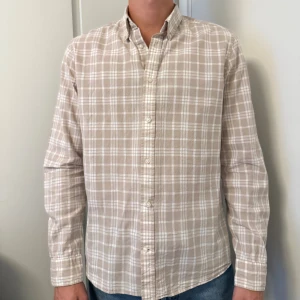 Beige rutig skjorta från Bondelid - Snygg beige skjorta från Bondelid i klassiskt rutigt mönster. Skjortan har långa ärmar, knappar framtill och en avslappnad passform. Perfekt för dig som gillar stilrena och tidlösa plagg. Tillverkad i mjuk bomull som känns skön mot huden.