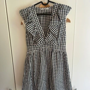 Nakd klänning - Supersöt kort klänning från NA-KD med svartvitt rutigt mönster, djup v-ringning och volangdetaljer vid axlarna. Klänningen är ärmlös och har en markerad midja som ger en fin siluett. Perfekt för dig som gillar en lekfull och trendig stil.