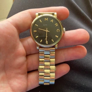 Marc by Marc Jacobs guld/silver klocka - Stilren klocka från Marc by Marc Jacobs med svart urtavla och guldiga detaljer. Armbandet är i rostfritt stål med både guld- och silverfärgade länkar. Snygg och modern design med tydliga bokstäver på urtavlan. Perfekt accessoar för att lyfta din outfit. Den behöver ett nytt batteri.