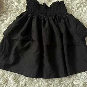 Svart volangkjol med smock - Supersöt svart kortkjol med bred smock i midjan och dubbla volanger. Kjolen har en luftig känsla och passar perfekt till sneakers eller boots. Enkel att styla med crop top eller hoodie för en chill look.