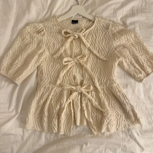 Krämvit blus med knyt från Gina Tricot - Supersöt krämvit blus från Gina Tricot i storlek XS. Blusen har puffiga halvlånga ärmar, strukturmönster i tyget och tre knyt framtill. Den har en lätt volang nertill, perfekt för dig som gillar detaljer och en feminin look.