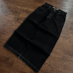 Svart jeanskjol från JNCO med brodyr - Svart lång jeanskjol från JNCO med kontrastsömmar, stora fickor bak och broderad snigel. Snygga randiga detaljer längs bak och rå kant nertill för en edgy vibe. Perfekt för dig som gillar streetstyle och unika plagg.