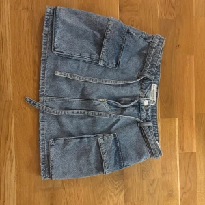 Blå jeanskjol med stora fickor - Säljer en blå jeanskjol från Stradivarius med stora fickor framtill och knytband i midjan. Kjolen har en klassisk denimlook och är perfekt för dig som gillar en avslappnad och trendig stil.