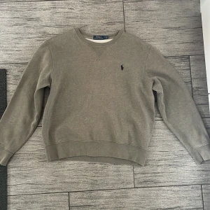 Grå sweatshirt från Polo Ralph Lauren - Säljer en stilren grå sweatshirt från Polo Ralph Lauren med klassisk rund halsringning och broderad logga på bröstet. Tröjan har långa ärmar och ribbade muddar för en skön passform. Perfekt för en chill och avslappnad look.