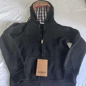 Burberry Kofta Svart - Svart hoodie från Burberry med klassiskt rutigt foder i huvan och guldfärgad dragkedja. Tröjan har snörning i huvan, ribbade muddar och känguruficka framtill. Tillverkad i mjuk bomull, perfekt för en clean och stilren look.