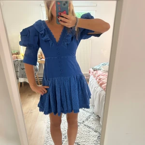 Blå kort klänning med volang och puffärm - Supersöt blå kort klänning med v-ringning, volangdetaljer och puffiga trekvartsärmar. Kjolen är utställd med broderade hål och knappar framtill. Perfekt för dig som gillar statement-ärmar och romantisk stil.