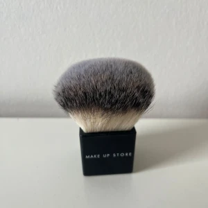 Kabuki-borste Make Up Store 409 - Kabuki-borste från Make Up Store, modell 409. Borsten har täta, mjuka syntetstrån i grått och vitt och ett stilrent svart fyrkantigt handtag. Perfekt för att applicera puder, bronzer eller rouge med jämnt och snyggt resultat.