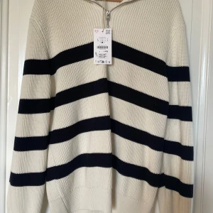 Randig stickad tröja med dragkedja - Säljer en krämvit stickad tröja med breda svarta ränder och halv dragkedja framtill. Tröjan har hög krage och långa ärmar, perfekt för en avslappnad och trendig look. Materialet är tjockt och mjukt, vilket gör den extra skön att bära.