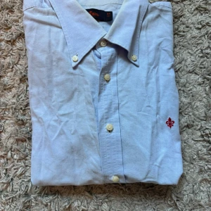 Ljusblå skjorta från Morris - Snygg ljusblå skjorta från Morris med klassisk krage och knappar framtill. Broderad röd lilja på bröstet ger en stilren touch. Perfekt för dig som gillar enkel men elegant stil.