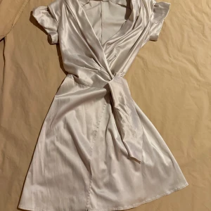 Champagnefärgad omlottklänning Smoph - Snygg kort omlottklänning från Smoph i champagnefärgad satin med knyt i midjan. Klänningen har korta ärmar, v-ringad hals och ett lyxigt, glansigt material som ger en elegant vibe. Perfekt för dig som gillar stilrena och trendiga plagg. Nypris 3000kr