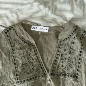 Olivgrön kort klänning från Zara - Supersöt olivgrön kort klänning från Zara med broderade detaljer. Klänningen har v-ringning, knappar framtill, långärmade puffärmar och markerad midja. Säljer då jag har två. Kan gå ner i pris 