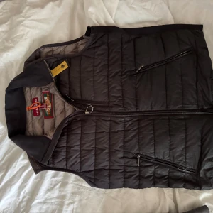 Svart quiltad dunväst Parajumpers S - Säljer en svart quiltad dunväst från Parajumpers i storlek S. Västen har dragkedja framtill, två snedställda fickor med dragkedja och en gul detalj vid kragen. Lätt och smidig modell med sportig känsla och snygg passform.