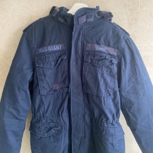 Mörkblå parkajacka från Brandit - Field jacket från Brandit i mörkblått med flera stora fickor och patchdetaljer. Jackan har ett mjukt teddyfoder på insidan som håller dig varm under kalla dagar. Klassisk militärinspirerad look med hög krage och dragkedja framtill.