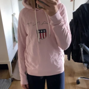 Ljusrosa hoodie från Gant  - Ljusrosa hoodie från Gant i storlek XS med klassisk logga och sköld framtill. Tröjan har huva med snörning och långa ärmar, har en brun fläck på ärmen men går nog att få bort i tvätten. 