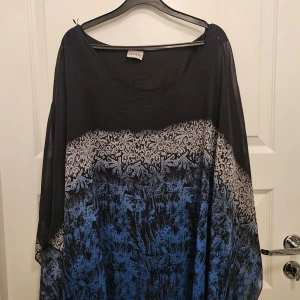 Festtunika från Emily i storlek 24/ EU 52 - Svart och blåmönstrad festtunika med strass från Emily. Tunikan är i ponchomodell och har ett svart linne under. Mycket gott skick.  Storlek 24/ EU 52 Bröstvidd 126 cm på linnet Längd 75 cm