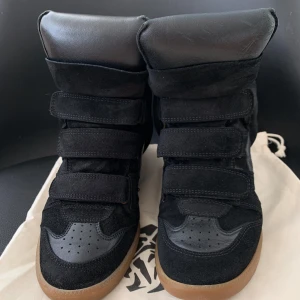 Isabel Marant svarta wedge sneakers - Svarta wedge sneakers från Isabel Marant med cool design och högt skaft. Skorna har kardborreband, detaljer i mocka och skinn samt gummisula i kontrasterande brun färg. Insidan är fodrad i ljust skinn för extra komfort. Perfekta för dig som vill ha en edgy streetstil.