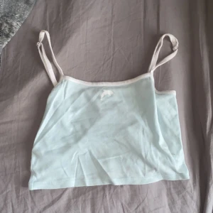 Ljusblå linnetopp från H&M Divided - Supersöt ljusblå linnetopp från H&M Divided med smala, justerbara axelband och vita kanter. Toppen är croppad och har en liten broderad delfin framtill. Perfekt för varma dagar och enkel att matcha med jeans eller kjol.