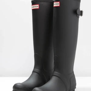 Svarta höga stövlar från Hunter - Klassiska svarta höga stövlar från Hunter med rund tå och justerbart spänne på sidan. Tillverkade i slitstark syntet som håller dig torr i regnet. Perfekta för blöta dagar och stilrena till alla outfits.
