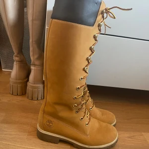 Timberland höga kängor i mocka - Snygga höga kängor från Timberland i klassisk ljusbrun mocka med snörning hela vägen upp och robust platt sula. Svart vadderad kant upptill för extra komfort. Perfekta för höst och vinter. Bokstavligt använda endast en gång.