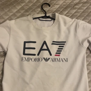 Vit EA7 Emporio Armani sweatshirt - Snygg vit sweatshirt från EA7 Emporio Armani med stor logga framtill i svart och rött. Klassisk rund halsringning och mjukt material som känns skönt mot huden. Perfekt för dig som gillar stilrena och trendiga plagg med tydlig branding.