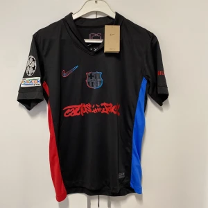 FC Barcelona x Nike Lamine Yamal tröja S - Säljer en ny oanvänd FC Barcelona matchtröja från Nike i svart med röda och blå detaljer, Lamine Yamal #19 på ryggen och coolt graffiti-tryck på framsidan. Kortärmad med Dri-FIT material som andas, officiella Champions League och UNHCR-loggor på ärmarna.