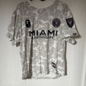 Miami BAPE x Inter Miami fotbollströja - Snygg fotbolls inspirerad tröja från BAPE x Inter Miami med grått och vitt camouflage-mönster. Tryck med MIAMI och BAPE 93 i svart, samt patchar på bröst och ärm. Rund hals och korta ärmar. Perfekt för dig som gillar streetwear och fotboll. Säljer då den va för liten