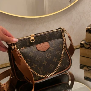 Louis Vuitton axelväska - Säljer en snygg axelväska från Louis Vuitton i klassiskt monogrammönster. Väskan är brun med ljusbruna detaljer, guldfärgad kedja och dragkedja. Justerbar axelrem i textil och läder, samt en liten rund myntplånbok som hänger på remmen. Perfekt för dig som gillar lyxiga accessoarer.