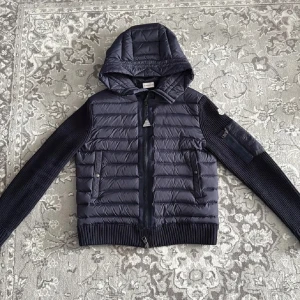 Mörkblå Moncler cardigan  - Snygg mörkblå cardigan från Moncler med stickade ärmar och vadderad kropp. Jackan har huva, dragkedja framtill och två fickor med tryckknappar. Moncler-logga på ena ärmen. 10/10 skick. Snabb affär = bättre pris 🤩