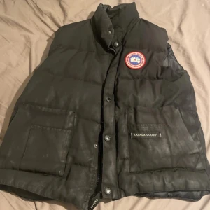 Svart dunväst från Canada Goose - Svart dunväst från Canada Goose med klassisk Arctic Program-patch på bröstet. Västen har två stora fickor framtill med logotyp, hög krage och både dragkedja och knappar. Materialet är glansigt och den har Synlig användning, perfekt för kalla dagar. Den är väldigt använd och saknas typ 2-3 knappar men den har fortfarande dragkedja