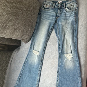 Blå bootcut jeans med slitningar - Snygga ljusblå bootcut jeans från gina med slitningar på knäna och coola röda detaljer på fickorna. Jeansen har klassisk femficksdesign, kontrastsömmar och breda benslut. Perfekt för dig som gillar en avslappnad och trendig look.