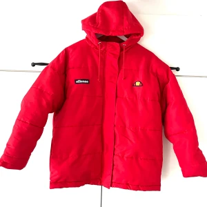 Röd pufferjacka från Ellesse - Röd pufferjacka med huva från Ellesse. Jackan har dragkedja och tryckknappar framtill, samt två logotyper: en på vänster bröst och en mindre på höger bröst.Jackan är tillverkad i 100% polyester vilket gör den mjuk och skön att bära. Två ytter fickor som håller händerna varma.passar perfekt att ha på höst och vinter. Passar Unisex , Barn och vuxna,ordpris:1599kr Bilderna togs personligen av mig, vilket säkerställer att du ser exakt den produkt du köper.vänligen observera att jag inte har öppetköp