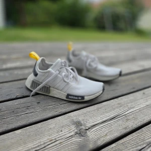 Adidas NMD-R1 ljusgrå sneakers med svarta/gula detaljer - Snygga Adidas NMD-R1 sneakers i ljusgrått och svarta detaljer samt en gul hälflik.. Ovandelen är i textil och sulan är vit med grå inslag. Klassisk tre-randig design på sidan och snörning framtill. Perfekta för dig som gillar streetstyle och bekväma skor.