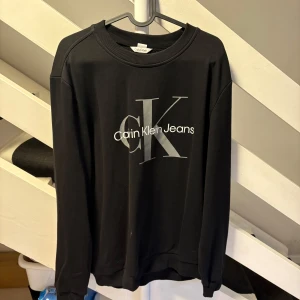 Svart Calvin Klein Jeans sweatshirt M - Svart sweatshirt från Calvin Klein Jeans i storlek M. Tröjan har en stor CK-logga och text på bröstet i grått och vitt. Klassisk rund halsringning och långa ärmar. Tillverkad i mjuk bomullsmix, perfekt för en clean och stilren look.