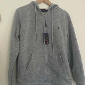 Grå hoodie från Polo Ralph Lauren - Snygg grå hoodie från Polo Ralph Lauren med dragkedja, huva och snörning. Klassisk logga broderad på bröstet och två fickor framtill. Säljer denna på grund av att jag vill ha en annan färg den är helt o använd där jag fick den som en present. Priset kan diskuteras och sänkas. Köpt 1200kr säljer 549kr