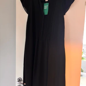 Svart klänning från H&M Conscious - Svart klänning från H&M Conscious med v-ringad hals och fina veckade detaljer framtill. Klänningen har korta ärmar och är tillverkad i ett lätt, flytande polyestertyg som ger en snygg siluett. Perfekt för dig som gillar stilren och enkel design.