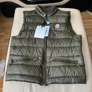 Olivgrön dunväst från Moncler - Snygg olivgrön dunväst från Moncler med quiltad design och två dragkedjeförsedda fickor. Västen har ståkrage, Moncler-logga på bröstet och smidig dragkedja framtill. Perfekt för lager-på-lager och chill stil under höst och vinter.
