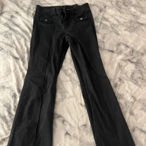 Svarta bootcut byxor med fickor framtil - Snygga svarta byxor med bootcut passform och raka ben. Framtill finns två fickor med lock och knappdetalj. Inga bakfickor 