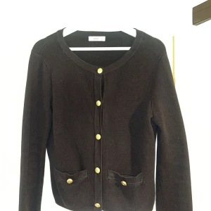 Svart kofta med guldknappar från Ellos - Stilren svart kofta från Ellos med rund halsringning och snygga guldknappar framtill. Två små fickor framtill med matchande knappar. S/M